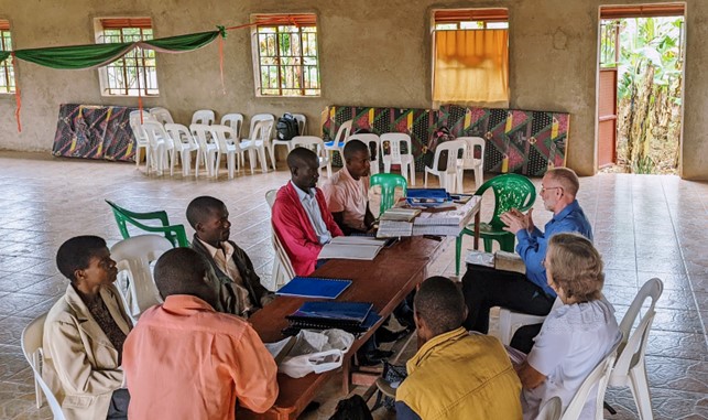 A group of Ugandan pastors sit at a long table with British pastors sat opposite. They are in conversation. There are plastic chairs and mattresses leant along a wall at the back