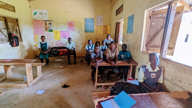 A few students in a run down classroom with missing desks. There are handdrawn posters on the walls and a rucksack hung on the window shutters